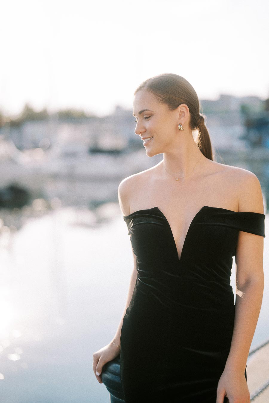 Woman in a black off-shoulder dress smiling beside a sunlit marina in Toronto.
