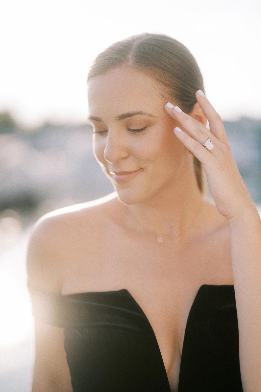 Bride-to-be smiles softly while showing her engagement ring in warm waterfront light.