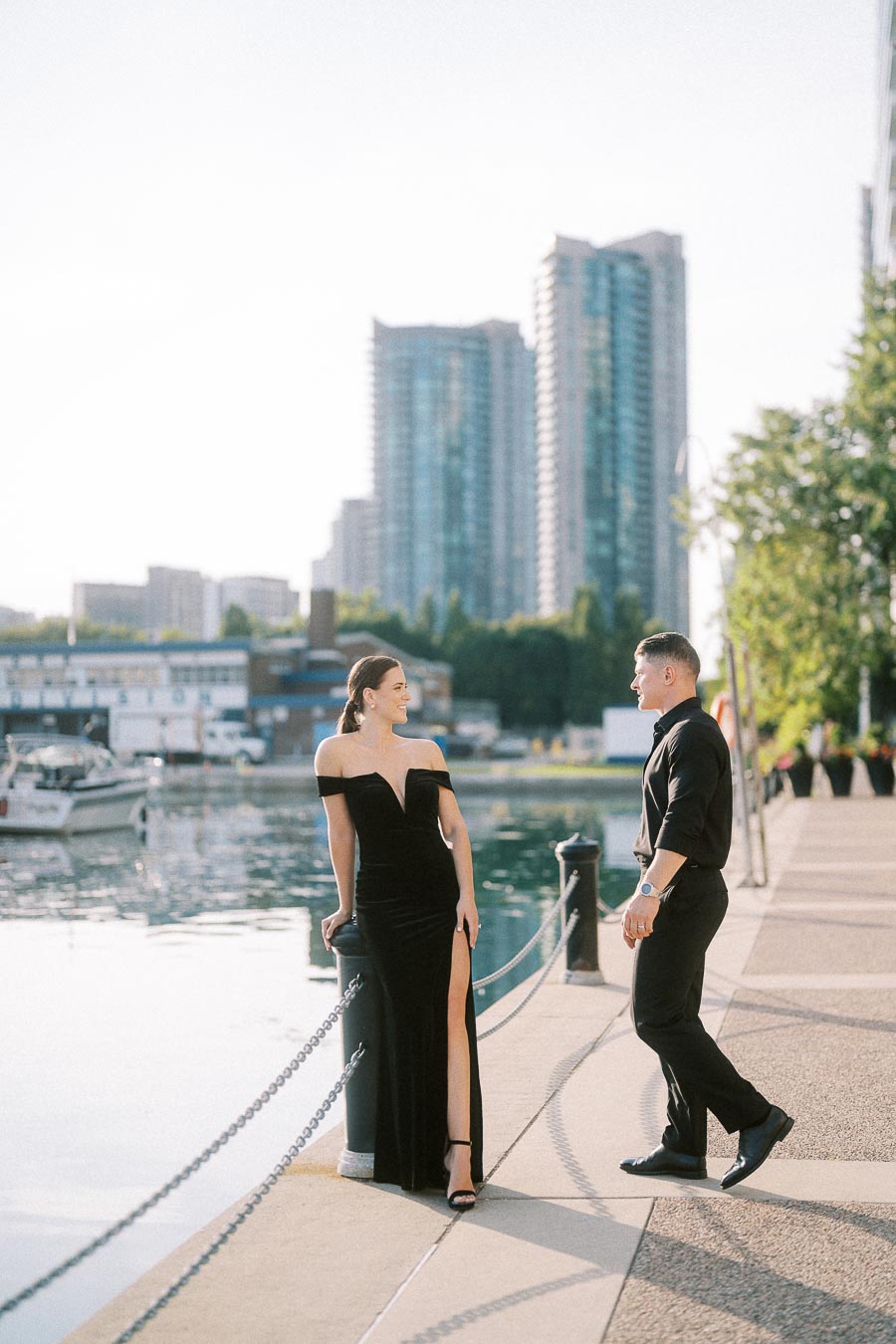 Couple in black formal outfits standing by a Toronto waterfront marina with high-rises behind them.