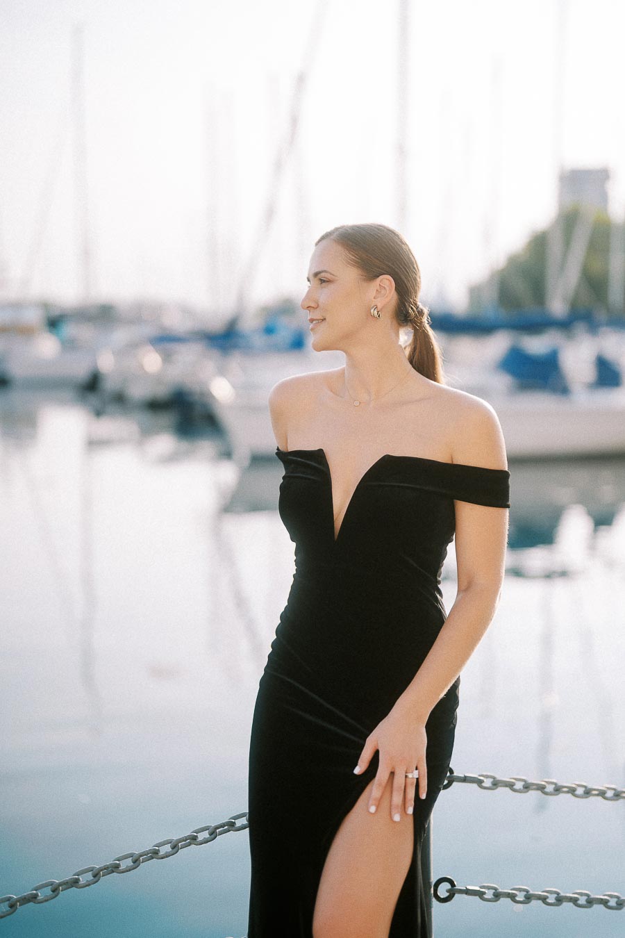 Woman in black off-shoulder dress stands by marina water with blurred sailboats behind her.