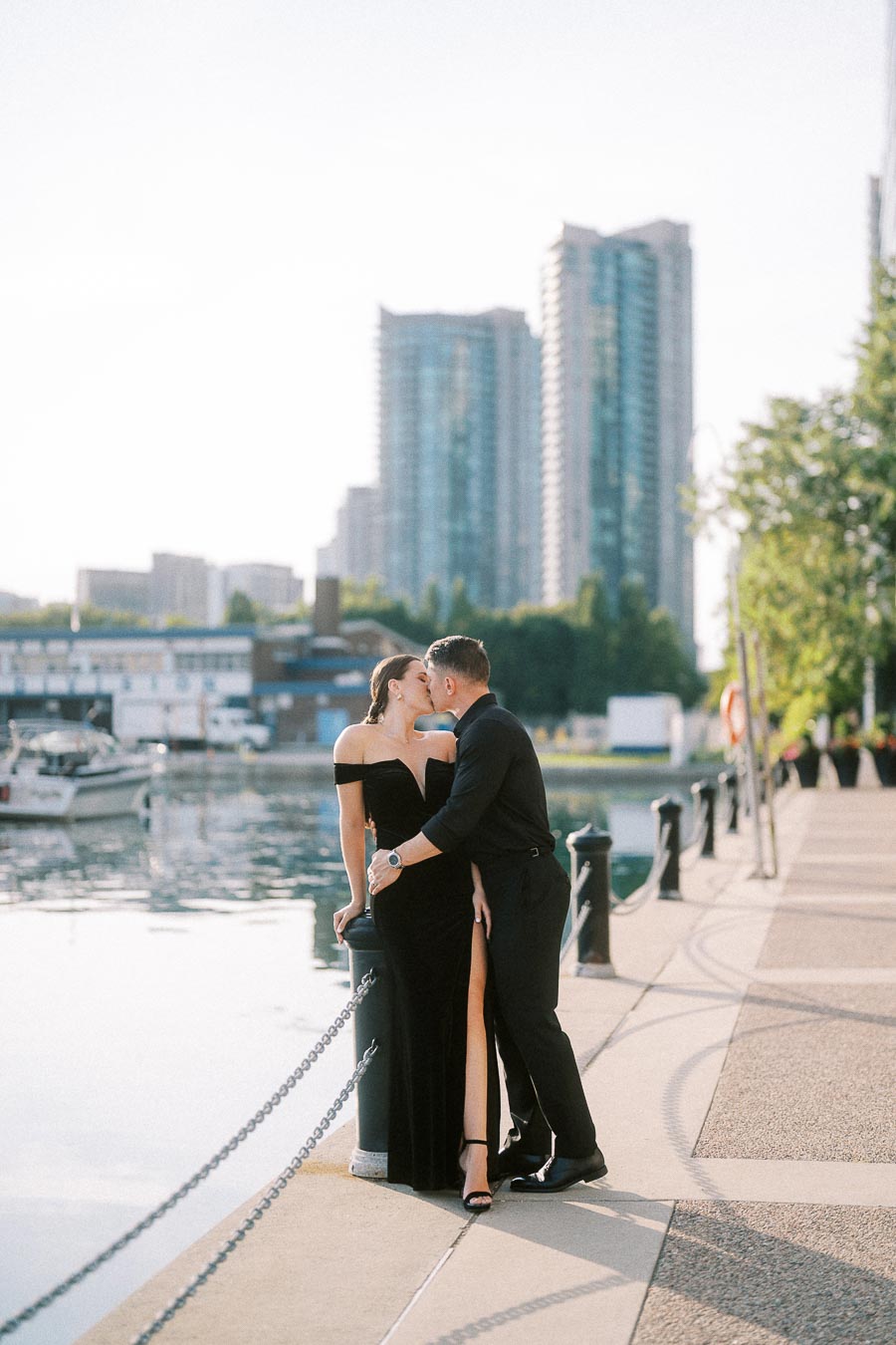 Couple in black formalwear kissing beside a Toronto waterfront marina with high-rise buildings behind them