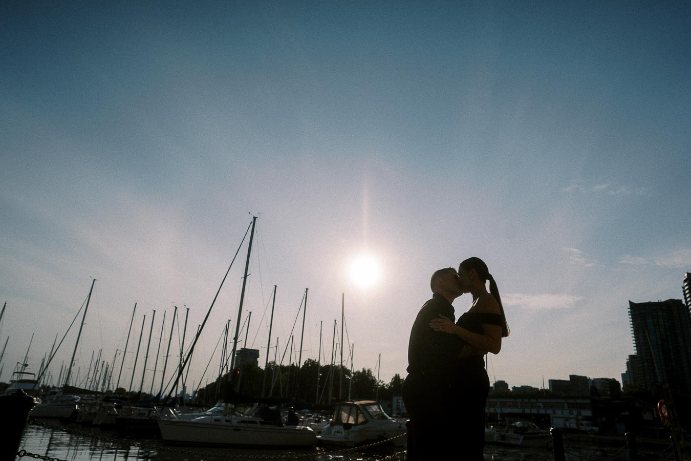 Silhouetted couple kissing at a Toronto waterfront marina, surrounded by sailboat masts at sunset.