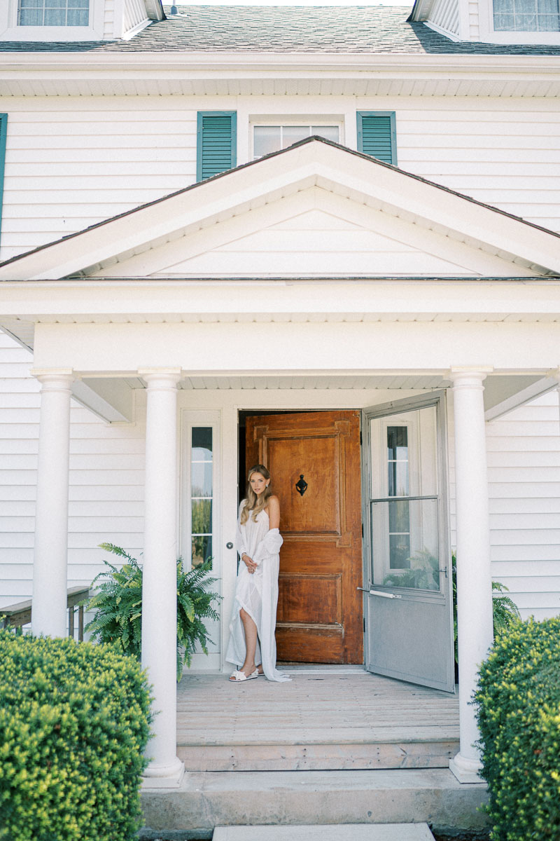 The bride stands at the farmhouse entrance at Martin Estates during a summer wedding in Niagara, Ontario.
