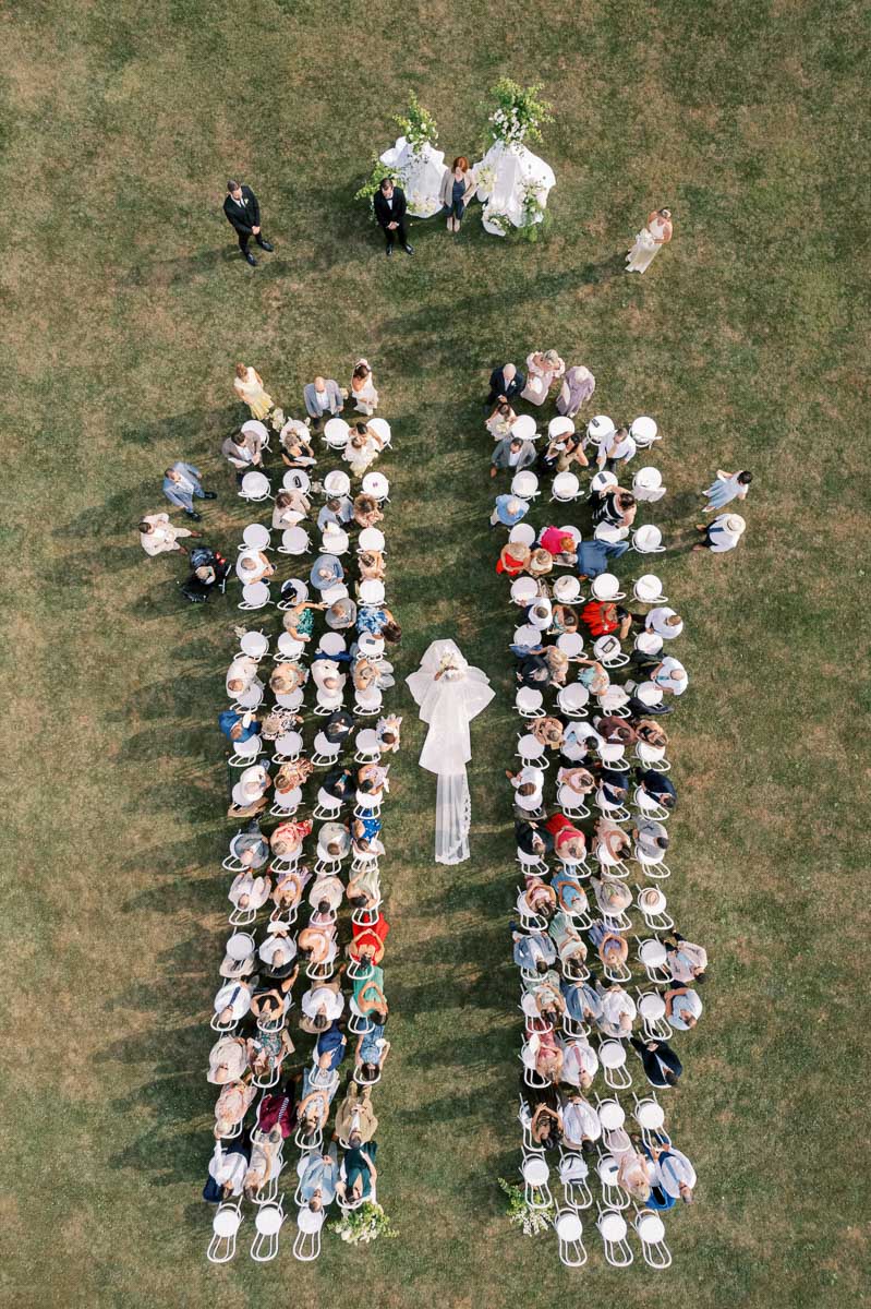 Aerial view of an outdoor wedding ceremony with a bride walking down the aisle between rows of seated guests on a grassy lawn, with an altar decorated with flowers in the background.